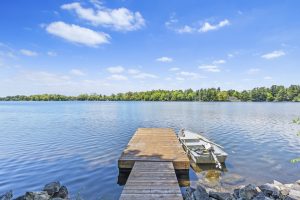 A peaceful rural lakefront view with a small wooden dock and motorboat, calm blue waters, and a tree-lined shoreline under a bright summer sky.