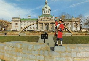 Kingston’s historic City Hall with its domed clock tower stands in the background, while Town Crier Chris Whyman, dressed in a red and blue uniform, raises his hand in proclamation at the front.