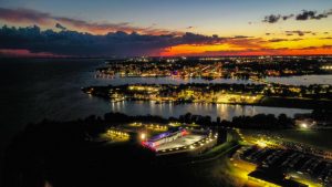 A dramatic aerial drone photo of Fort Henry illuminated at night during the Sunset Ceremony, with downtown Kingston and Lake Ontario glowing in the distance