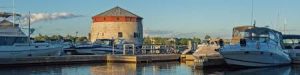 Shoal Tower, a round limestone Martello tower with a red roof, stands on Kingston’s waterfront behind a marina filled with docked boats.