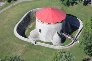 A clear aerial photo of Murney Tower in Kingston, Ontario, showing the round limestone Martello tower with its red roof, surrounding stone walls, and grassy parkland.
