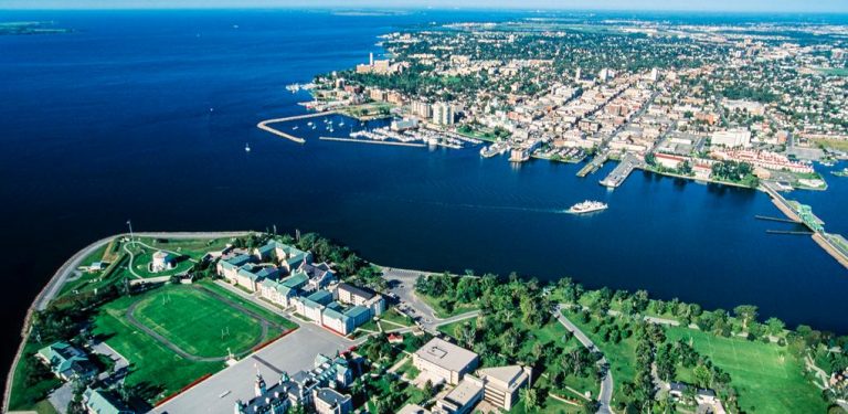 Aerial photo of Kingston, Ontario, showing the downtown waterfront, marinas, ferry on Lake Ontario, and surrounding cityscape.