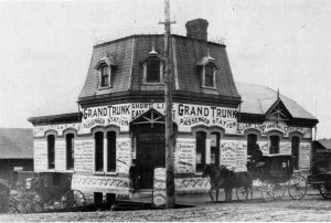 Historic photograph of the Grand Trunk Railway station at Ontario and Johnson Streets in Kingston, Ontario, taken in the 1890s. The station features a limestone foundation with brick walls, with horse-drawn carriages in front.