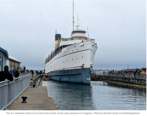 The S.S. Keewatin, a historic passenger steamship, docking at its new home at the Great Lakes Museum in Kingston, Ontario, with crowds gathered along the pier.